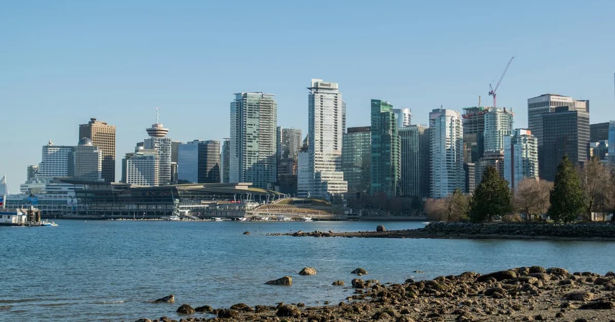 Downtown Vancouver skyline and the Vancouver Convention Centre seen across Coal Harbour from a rocky Stanley Park shoreline under a clear blue sky